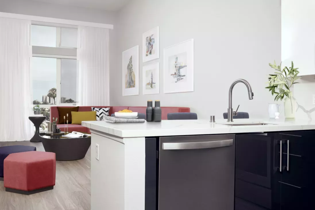 Modern kitchen island with stainless steel appliances and a gooseneck faucet, opening into a stylish living area with a red sectional, geometric ottomans, and framed wall art at The Linden.