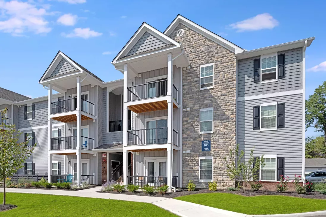 Three-story apartment building with gabled roofs, stone and siding exterior, and balconies overlooking the landscaped grounds.