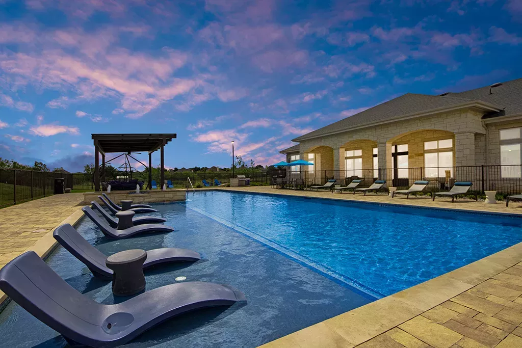 Luxury outdoor swimming pool at twilight with in-water lounge chairs, poolside seating, and clubhouse at 32Hundred Windsor Apartments in Denton, TX