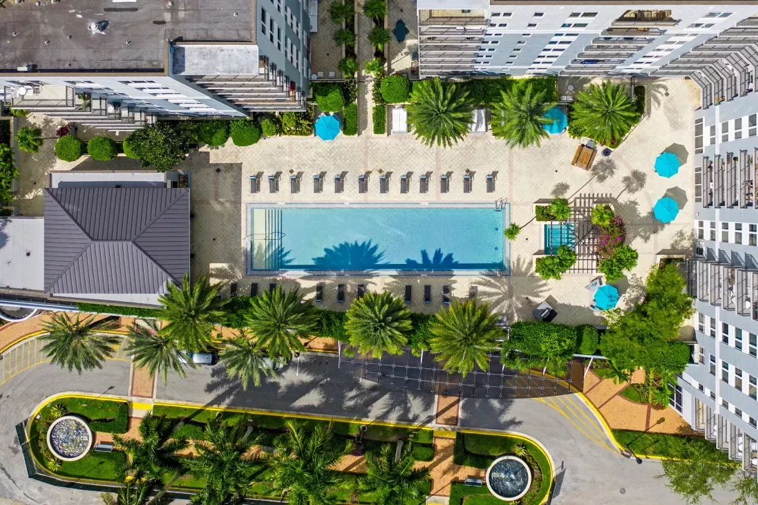 Aerial view of a luxury apartment courtyard at Midtown 24 in Plantation, Florida, featuring a rectangular swimming pool surrounded by lounge chairs, palm trees, and turquoise umbrellas. The landscaped area includes shaded seating, decorative walkways, and a small spa, creating a tropical resort-style atmosphere.