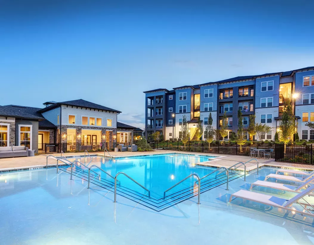 Resort-style outdoor swimming pool with lounge chairs, modern clubhouse, and apartment community at dusk.