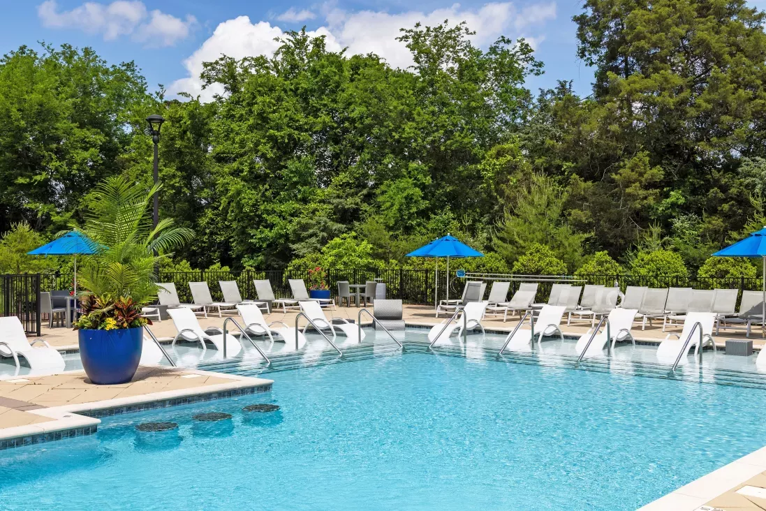 Resort-style swimming pool with in-water loungers, poolside chairs, and blue umbrellas surrounded by lush greenery.