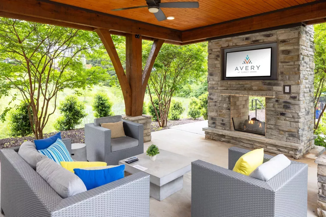 Covered outdoor lounge featuring modern wicker seating, colorful accent pillows, a stone fireplace with mounted TV, and views of landscaped greenery.
