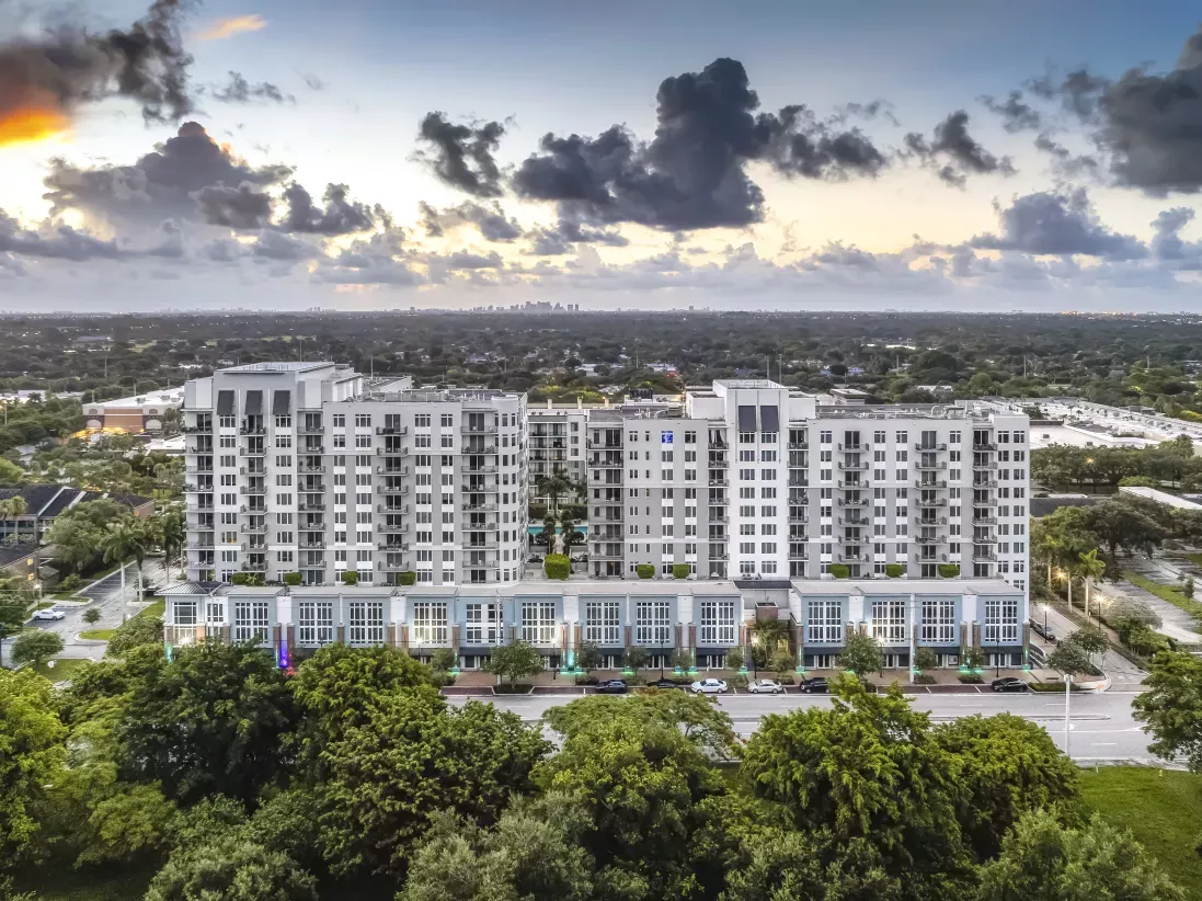 Aerial view of Midtown 24, a modern multi-story apartment complex in Plantation, Florida, featuring white and gray exteriors, private balconies, and ground-level retail spaces. Surrounded by lush greenery and city infrastructure, with the Fort Lauderdale skyline visible in the distant background under a partly cloudy sunset sky.