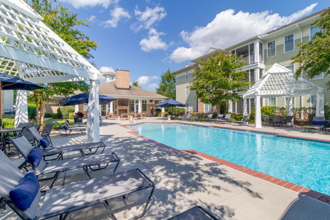 Resort-style pool area with lounge chairs, white pergolas, and a gazebo, surrounded by apartment buildings under a blue sky.