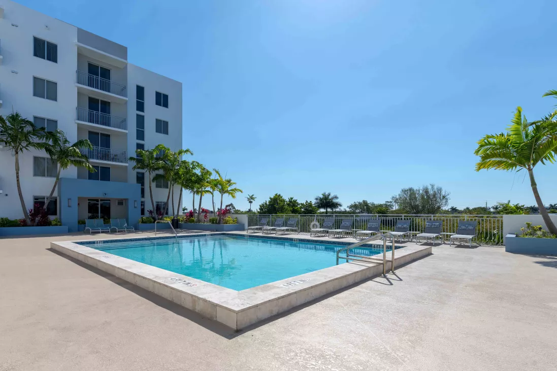 Bright swimming pool with lounge chairs and palm trees next to a modern apartment building under a blue sky.