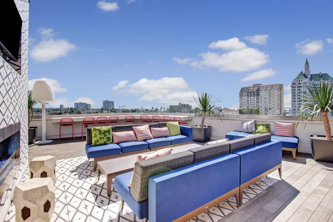 Sunny rooftop patio with modern blue couches, colorful pillows, and a bar overlooking a city skyline.