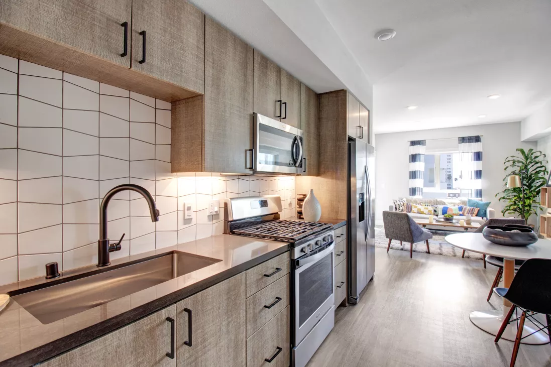 Modern kitchen features light wood cabinets, white tiled backsplash, stainless steel appliances. Open living room beyond.
