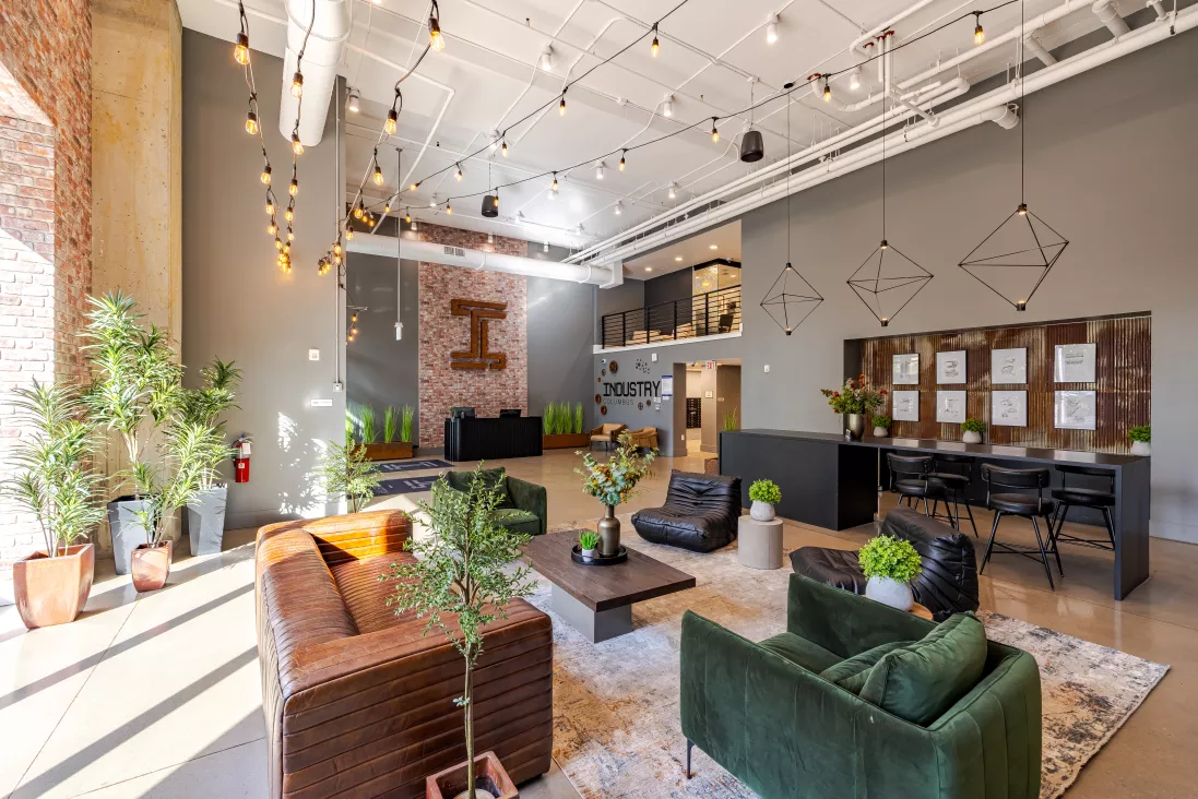 Modern industrial lobby with high ceilings, exposed bulbs, green and brown lounge chairs, reception desks, and potted plants.