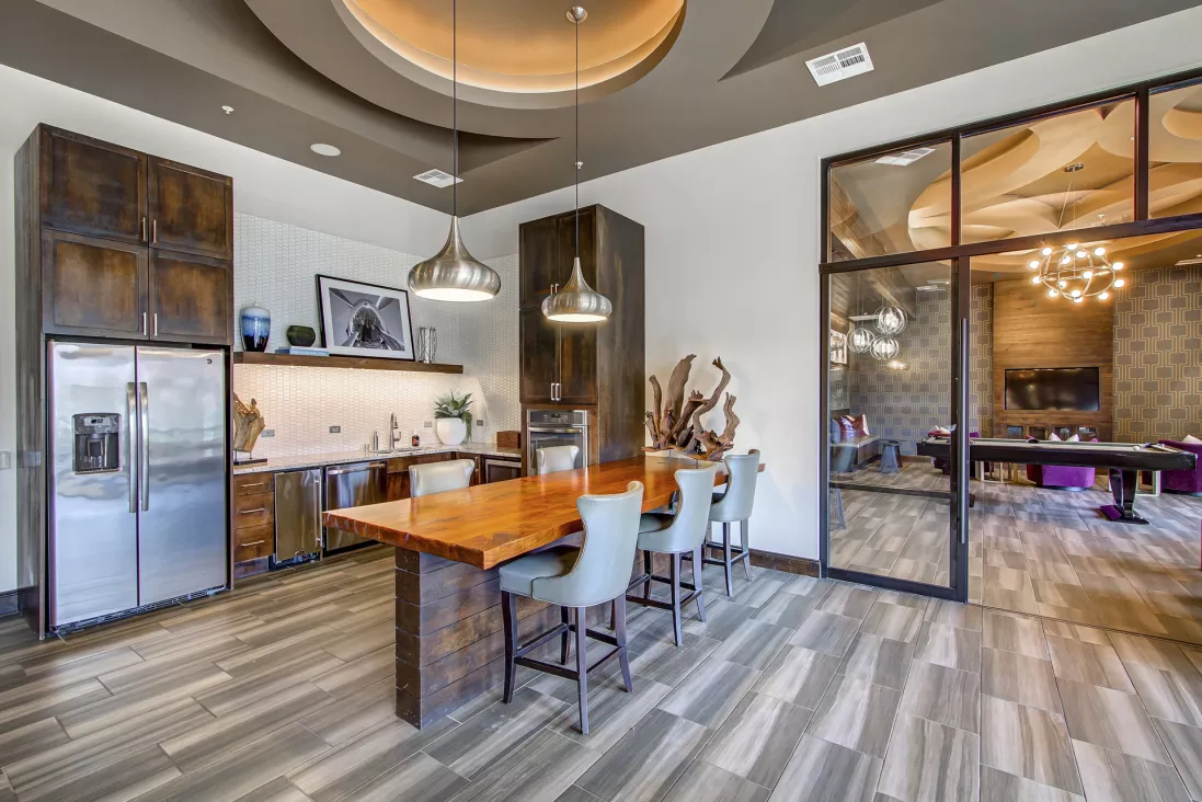 Modern apartment lounge with kitchen, communal wood table, and glass wall to a game room with pool table.