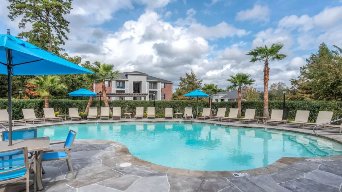 Outdoor swimming pool with lounge chairs, blue umbrellas, palm trees, and an apartment building under a partly cloudy sky.
