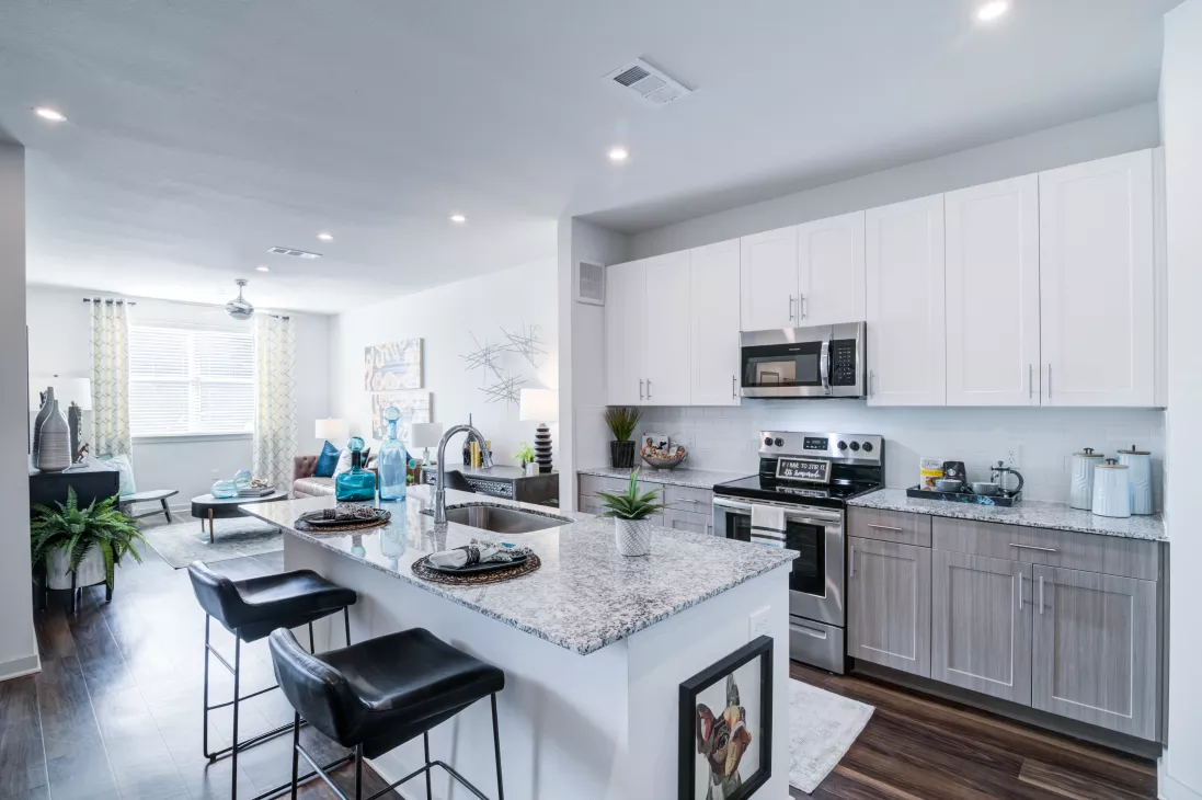 Modern open-plan kitchen and living room with white cabinets, stainless steel appliances, a granite island, and dark wood floors.