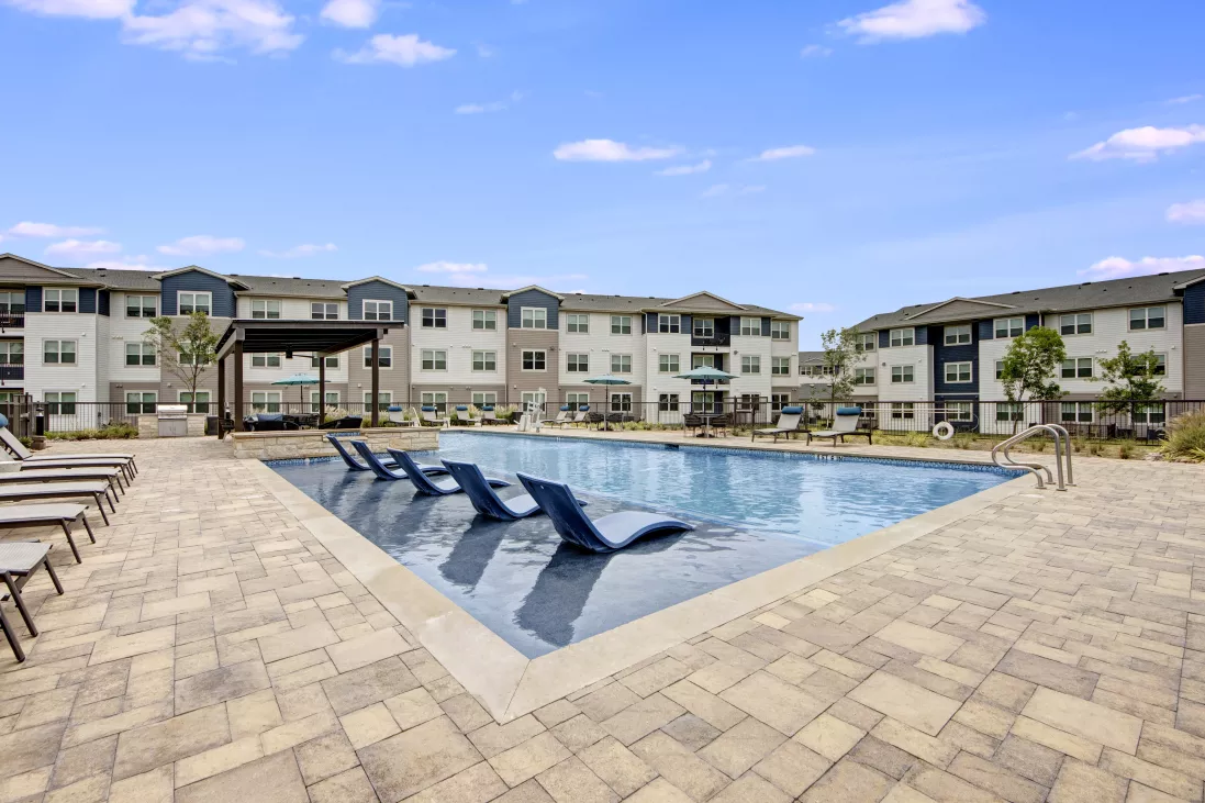 Outdoor swimming pool area with blue lounge chairs, surrounded by paved patio and modern apartment buildings.