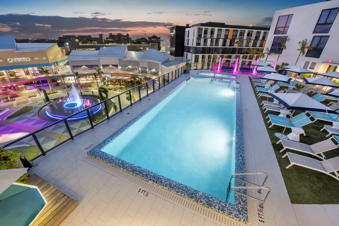 Photograph: Modern rooftop pool and lounge area glowing with blue and pink lights at dusk, overlooking a lively plaza.