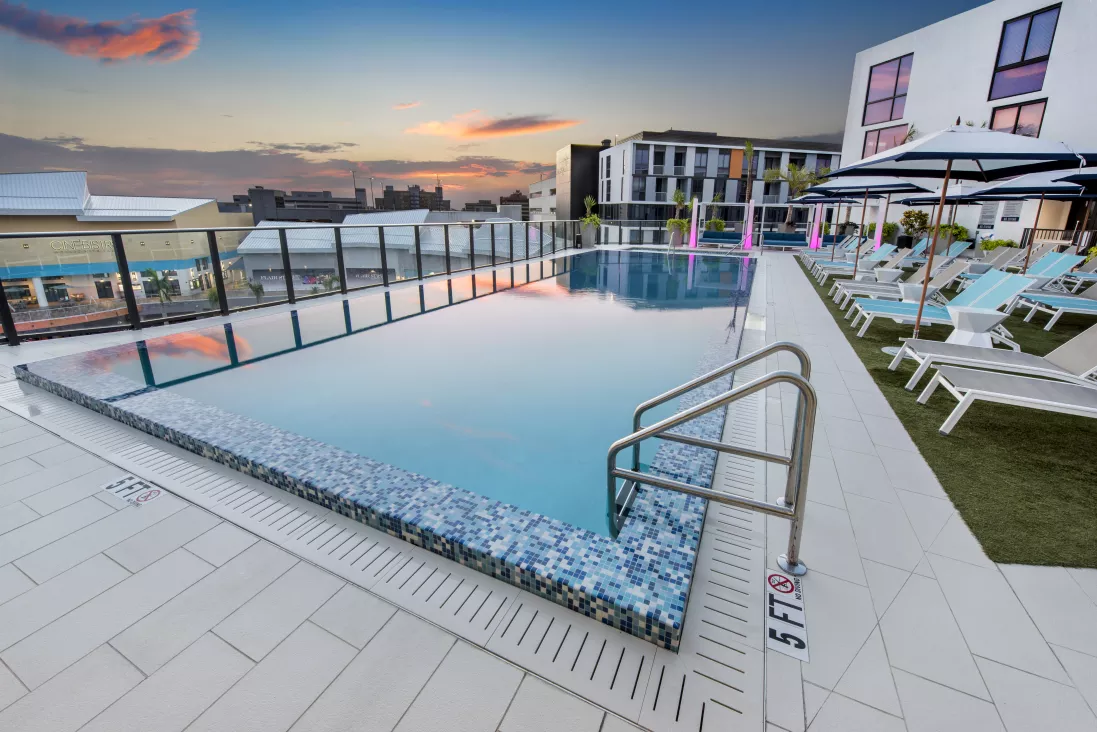 Rooftop infinity pool with blue mosaic tiles, lounge chairs, and modern buildings under a colorful sunset sky.