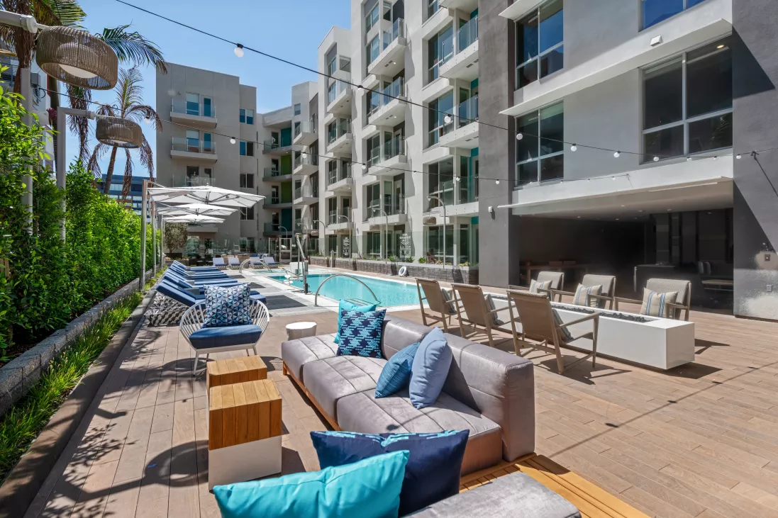 Modern apartment complex pool area with lounge chairs, sofas, and string lights on a sunny day. Tall buildings behind.