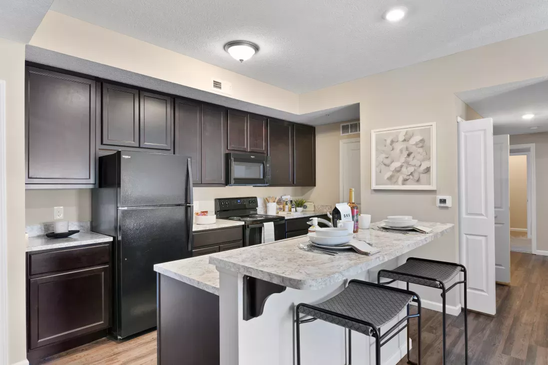 Modern kitchen with dark brown cabinets, black appliances, light speckled countertops, and a breakfast bar with two stools.
