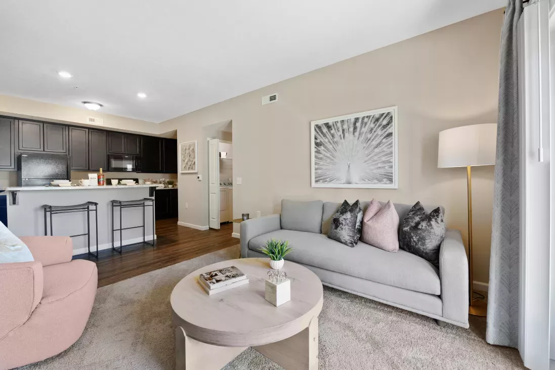 Modern open living room with gray sofa, pink armchair, wood coffee table, peacock art, and dark kitchen with breakfast bar.