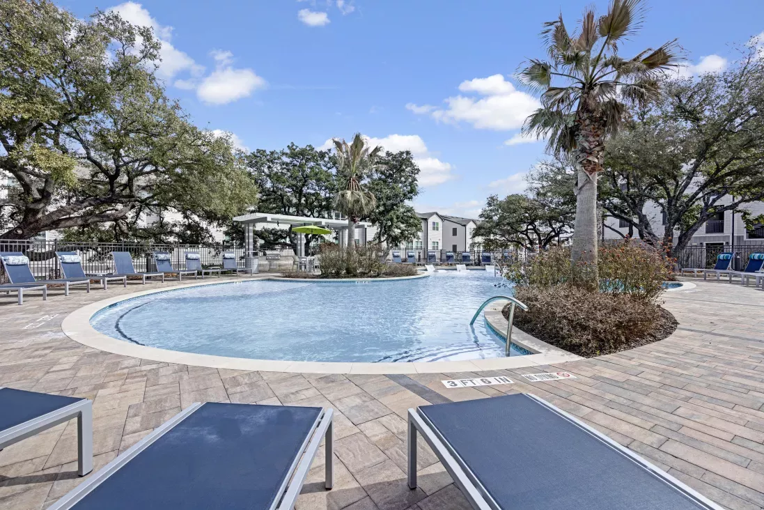 Large swimming pool with blue lounge chairs on a patterned patio, surrounded by trees and residential buildings under a blue sky.