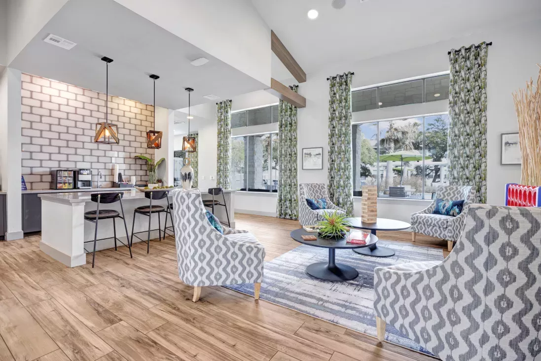 Modern communal lounge with a coffee bar, white brick wall, patterned armchairs, round coffee table, and large windows.