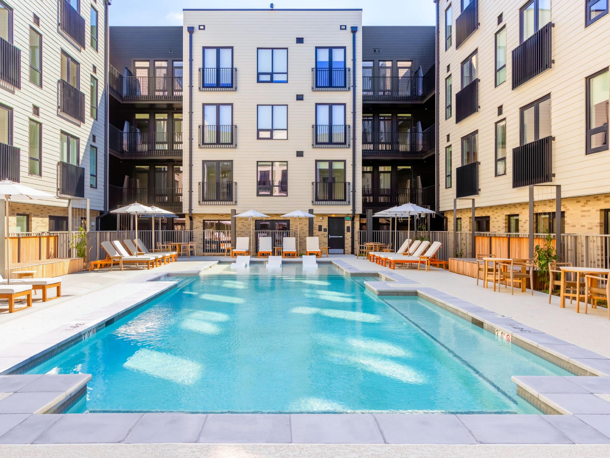 Outdoor pool area at The Edmund Apartments featuring lounge chairs, umbrellas, and in-pool seating surrounded by modern apartment buildings.
