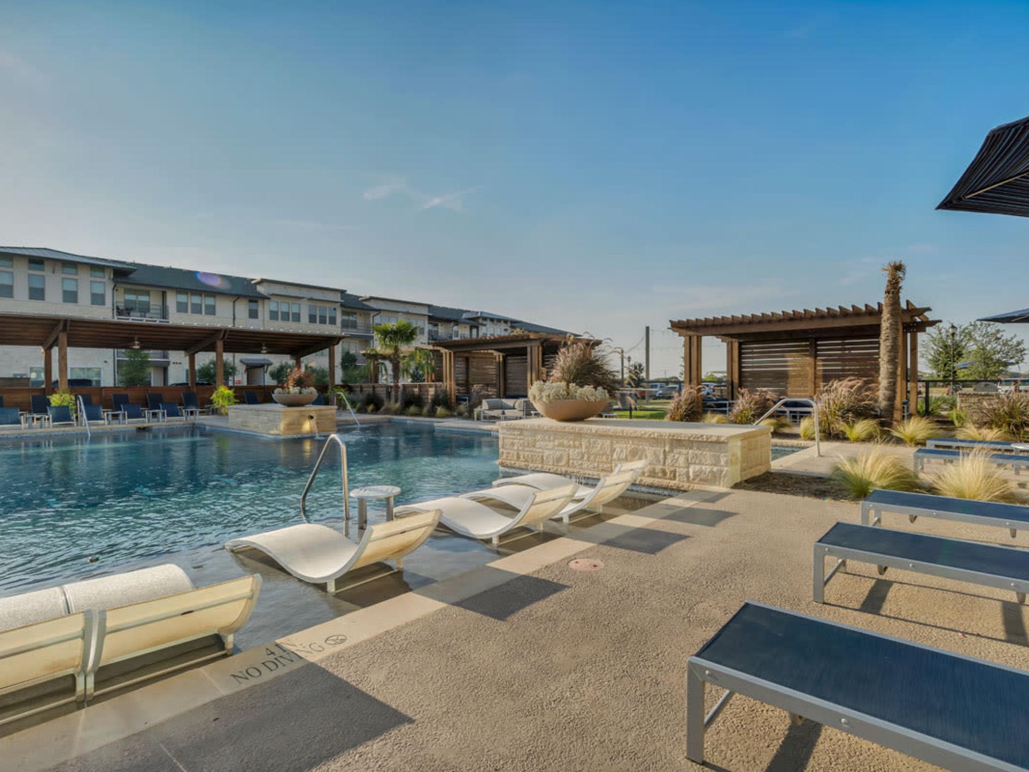A serene outdoor pool with sun loungers and shaded pergolas, surrounded by landscaping.