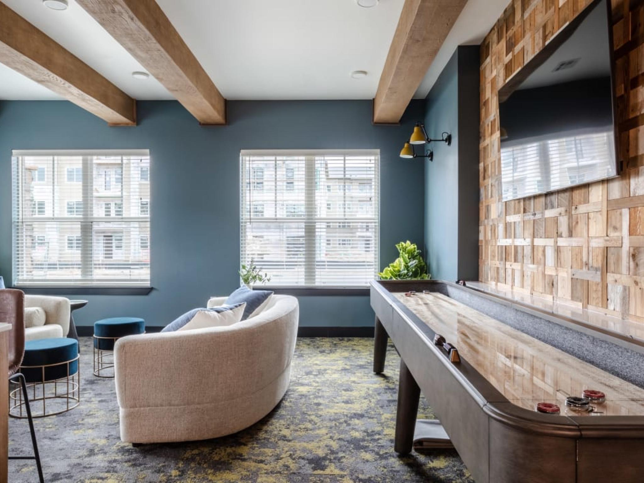 A stylish lounge area with a shuffleboard table, cozy seating, wooden accent walls, and large windows allowing ample sunlight.
