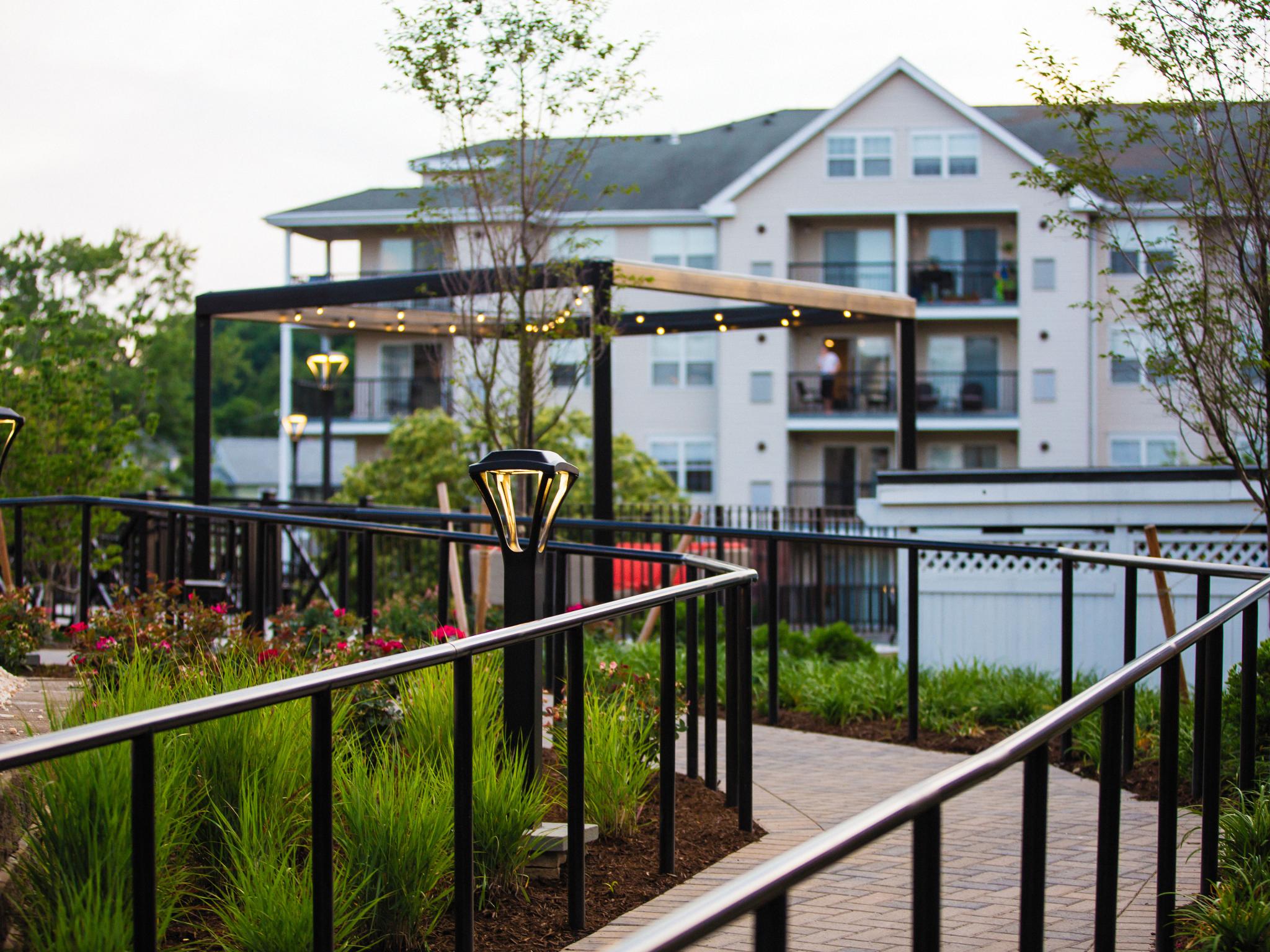 A beautifully landscaped walkway with modern railing, warm lighting, and lush greenery leading to an outdoor seating area.