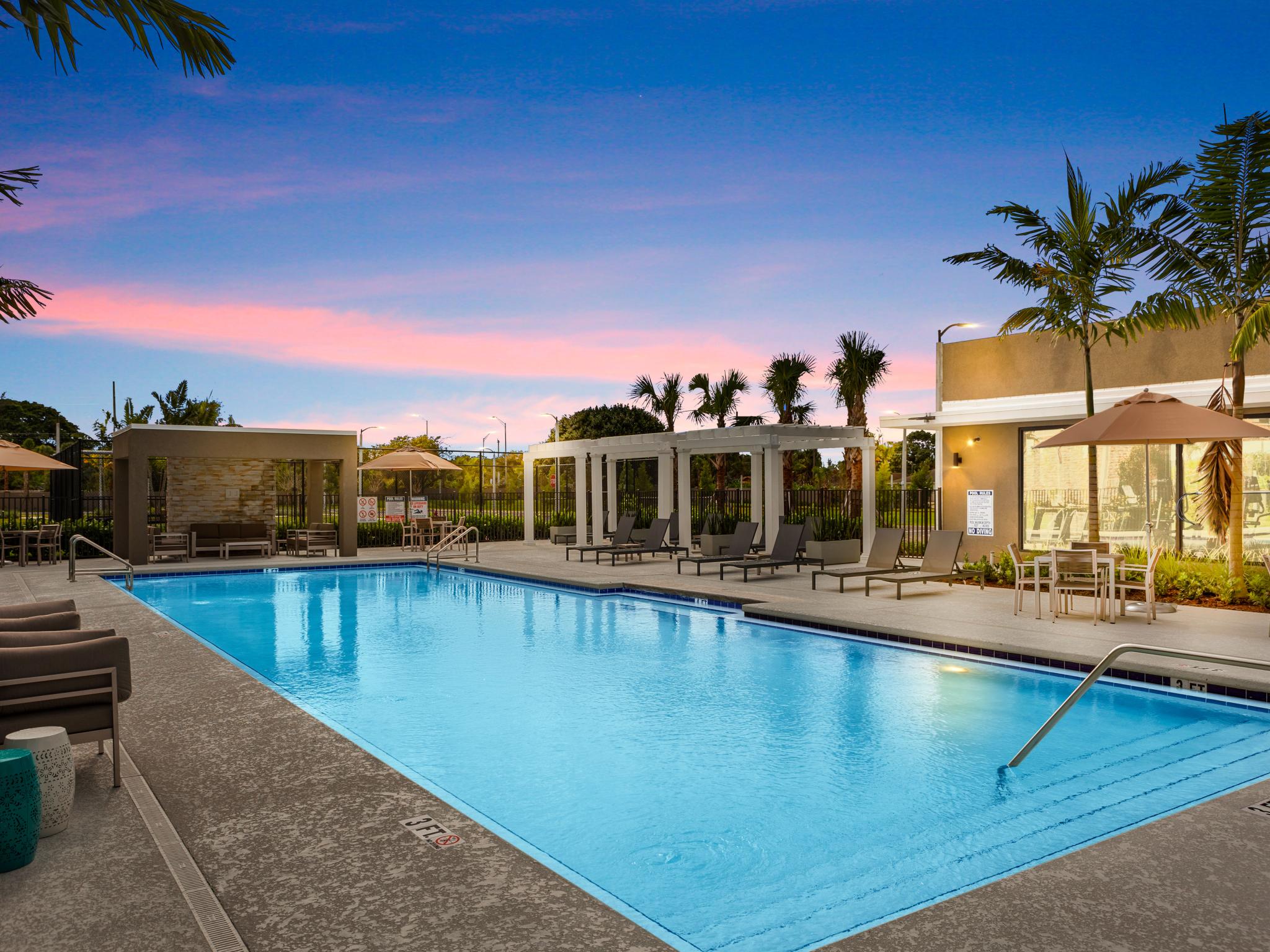 A beautifully lit outdoor swimming pool area at sunset, complete with lounge chairs, cabanas, and lush palm trees for a resort-style experience.