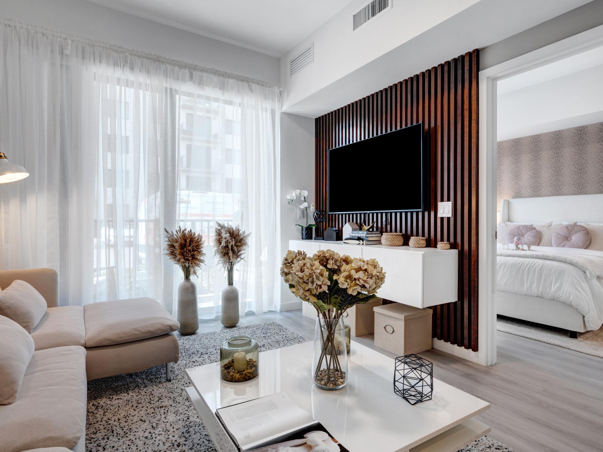 Bright and stylish living room with contemporary decor, large windows with sheer curtains, and a connected view of the bedroom.
