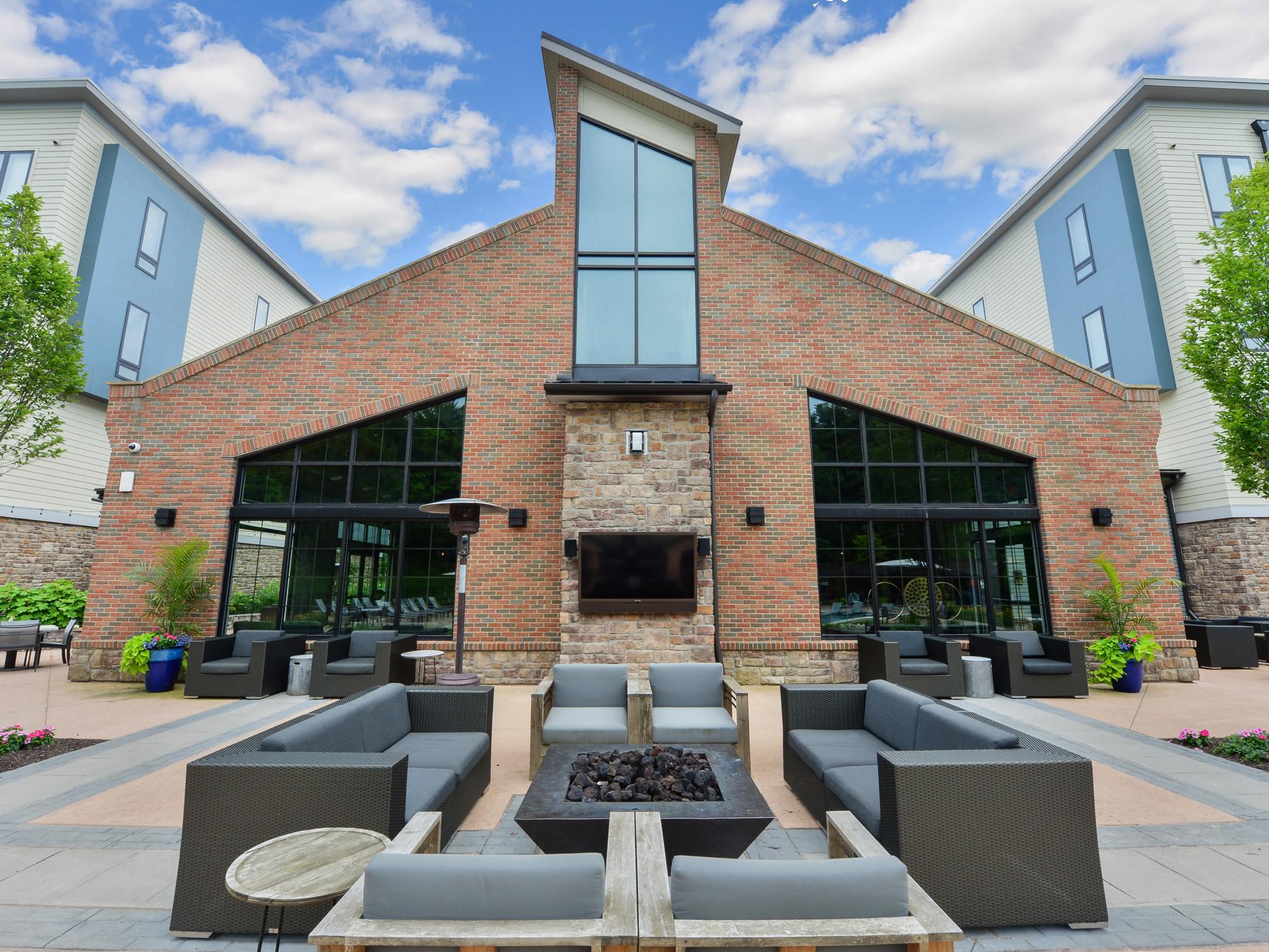 Modern outdoor lounge area featuring a brick and stone clubhouse facade, cozy fire pit, and stylish gray seating surrounded by lush greenery.