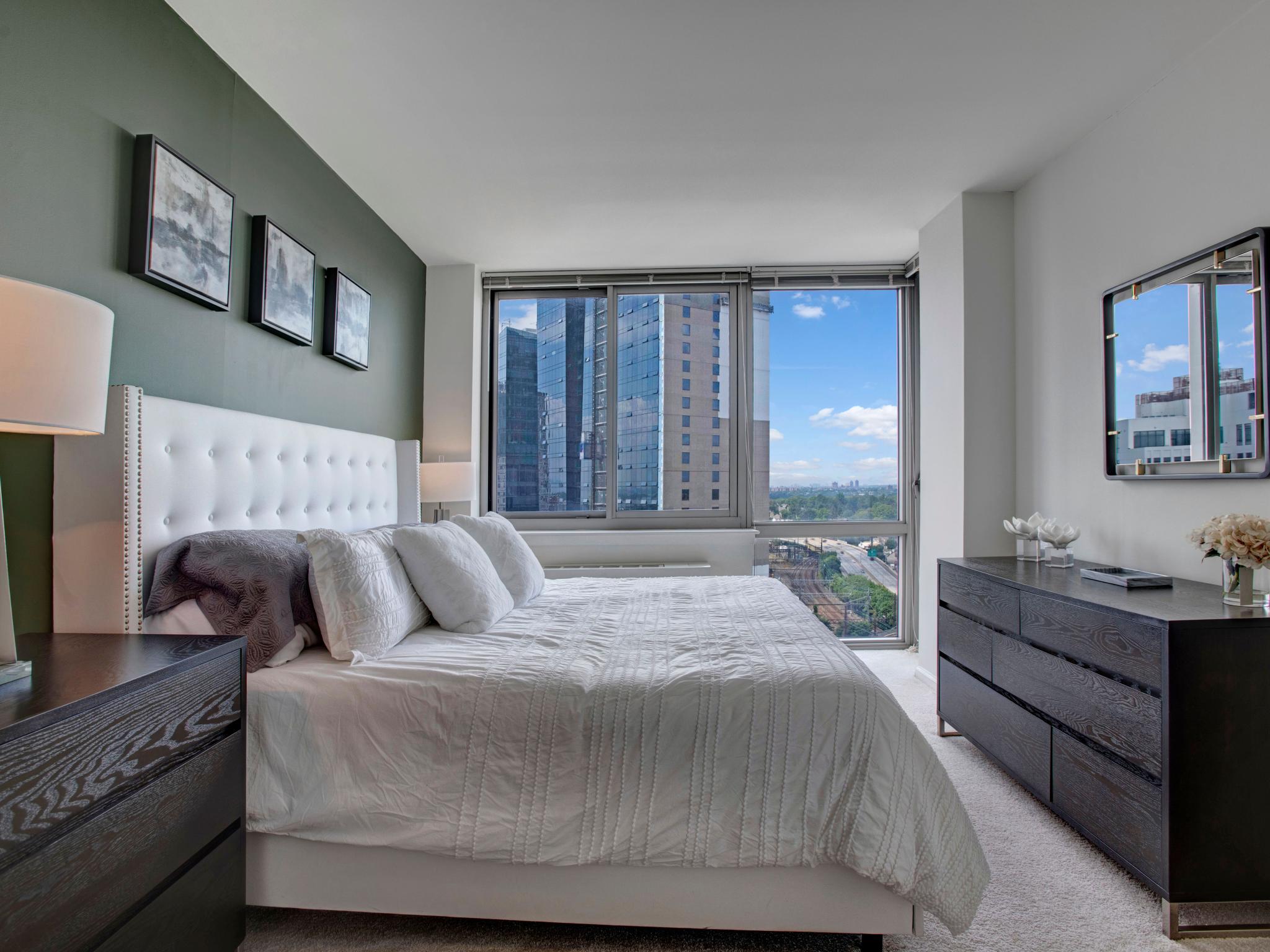Luxurious bedroom at Skyline New Rochelle with a tufted headboard, elegant furnishings, and large windows showcasing scenic urban vistas.