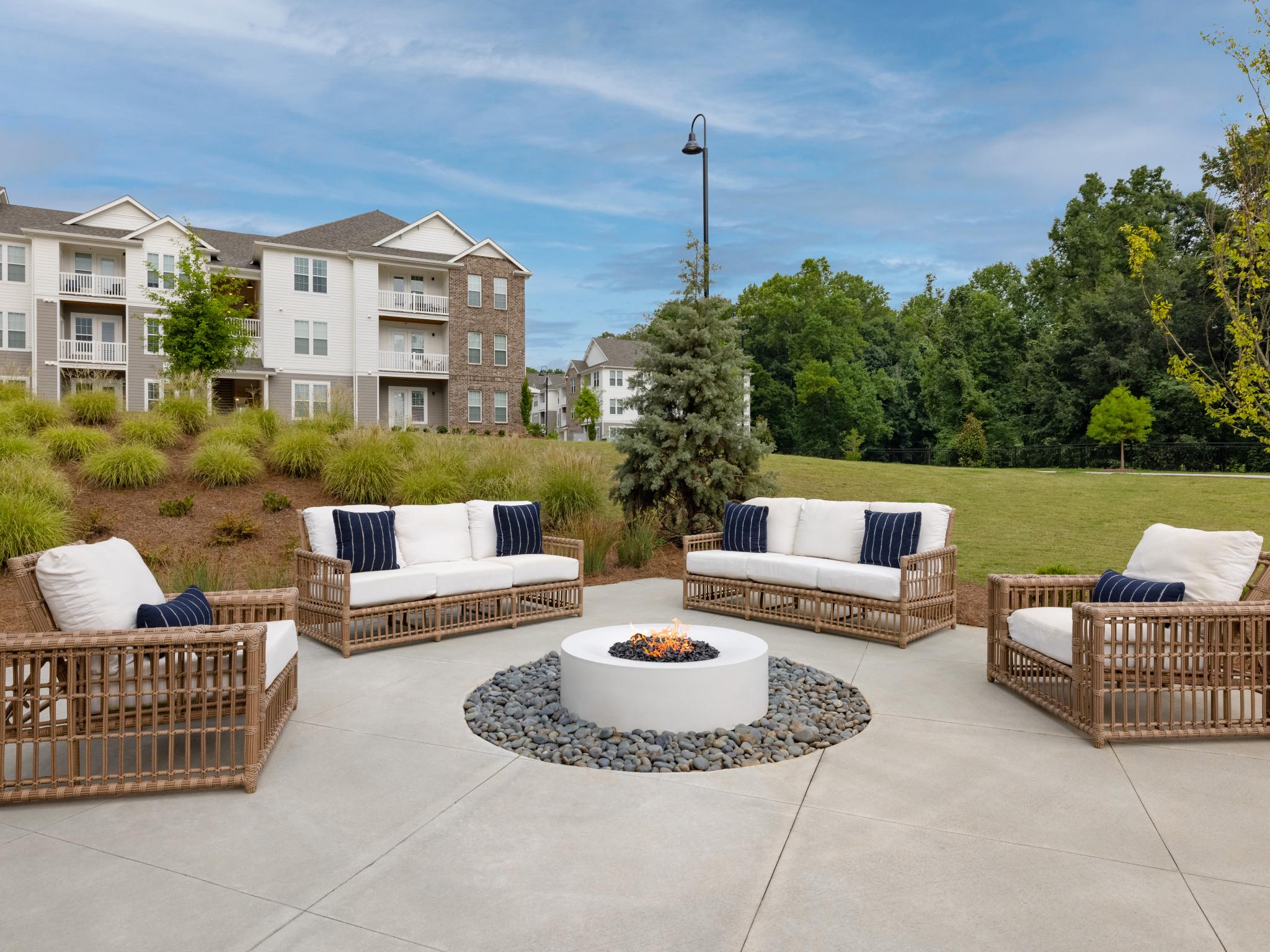 A stylish outdoor fire pit lounge featuring plush wicker seating with white cushions and navy accent pillows, surrounded by lush landscaping and modern apartment buildings in the background.