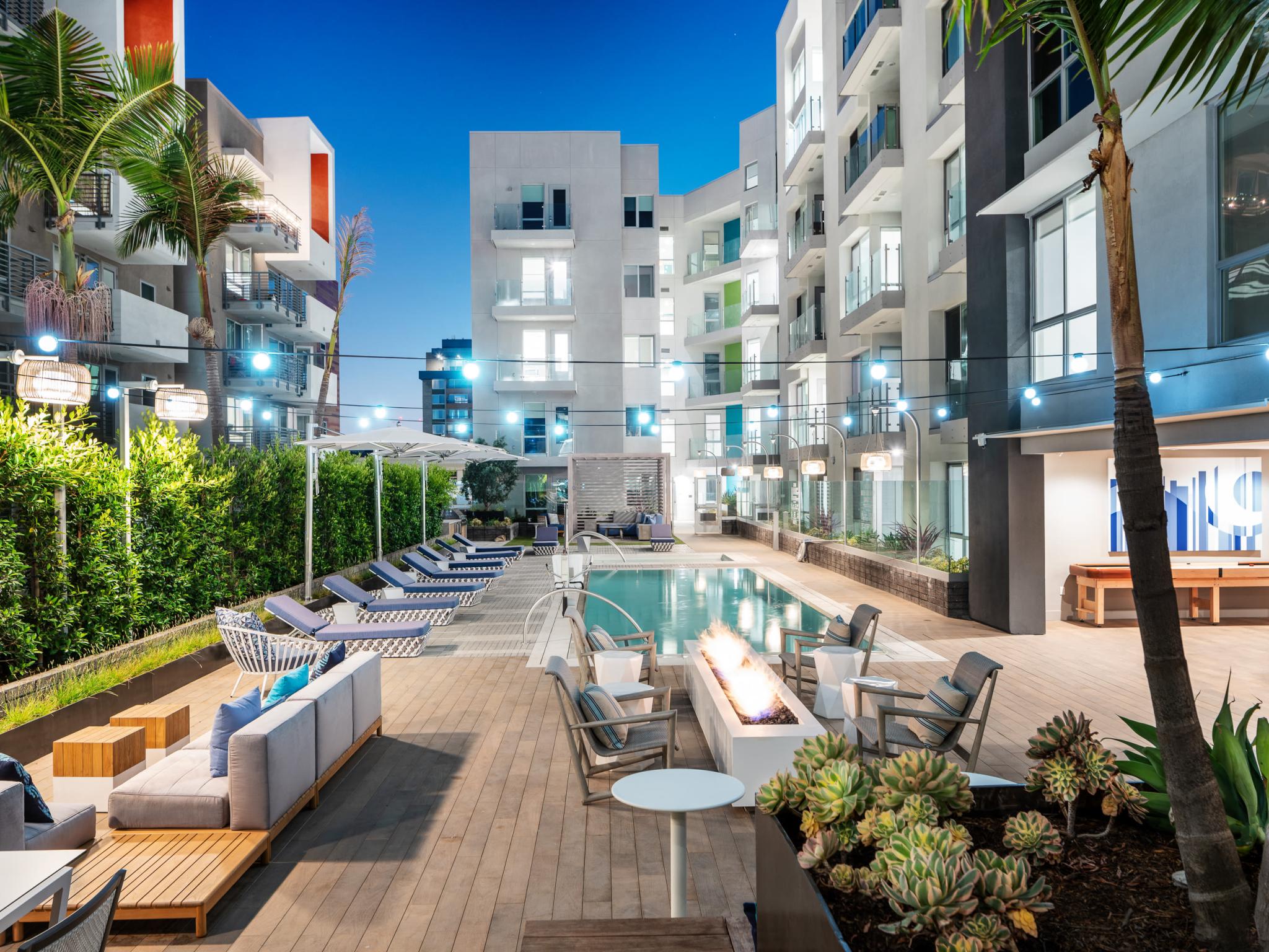 Resort-style pool area at The Linden Luxury Apartments showcasing lounge seating, ambient lighting, and a beautiful fire pit surrounded by lush greenery.