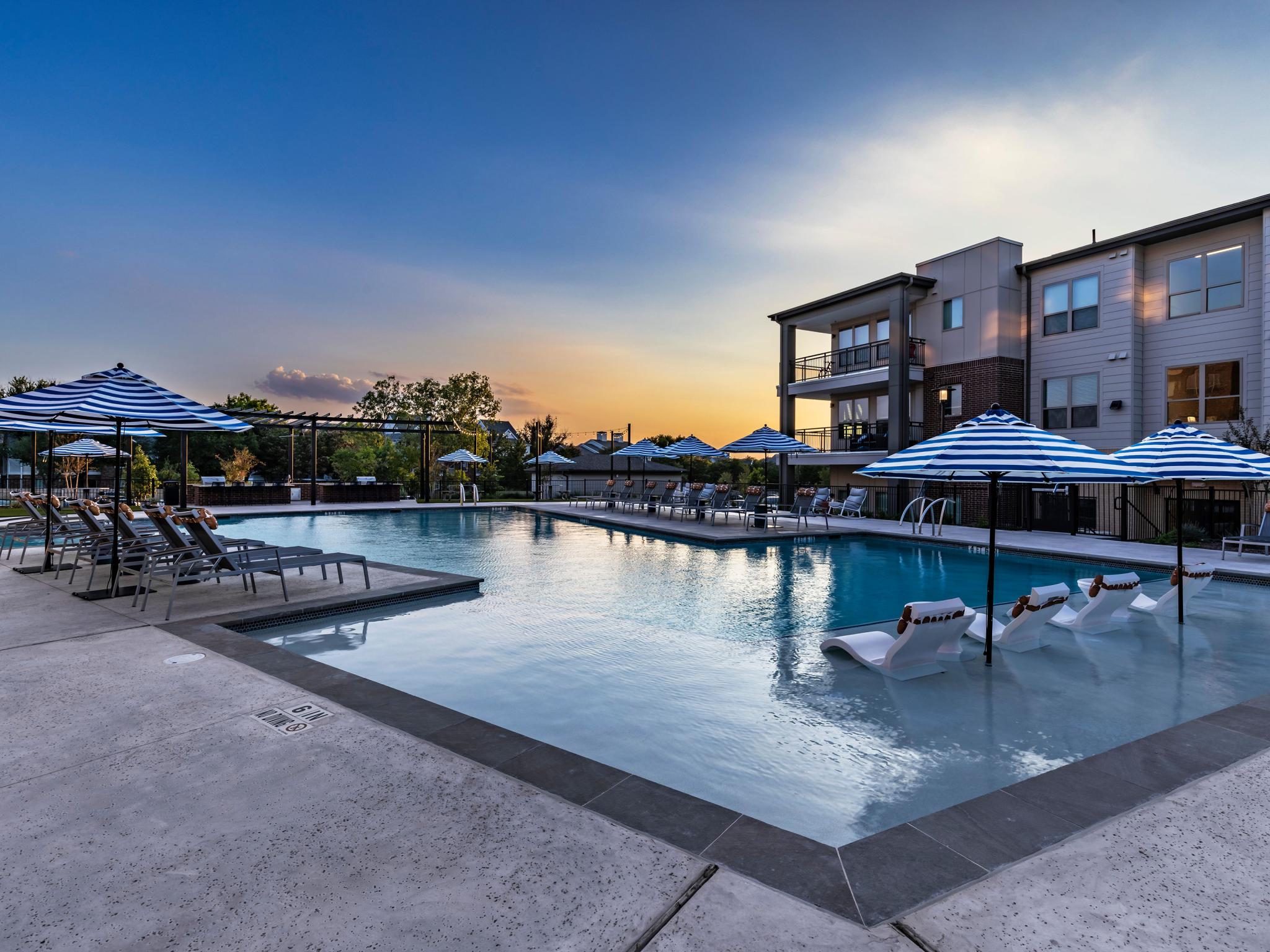 A breathtaking view of The Warner Apartments' pool at sunset, with in-water loungers, striped umbrellas, and elegant lighting creating a serene ambiance.