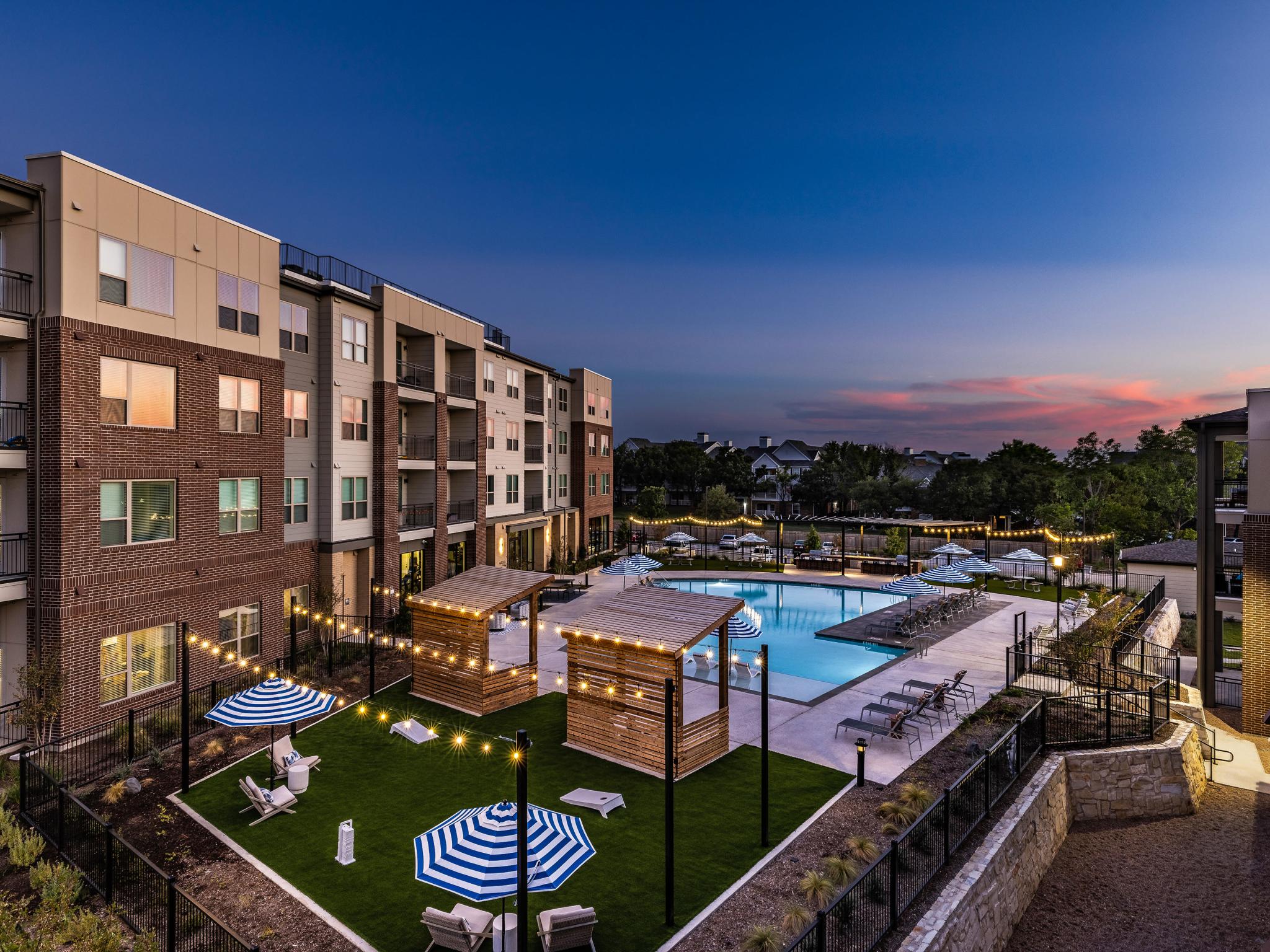 An aerial view of The Warner Apartments' pool and courtyard at dusk, featuring glowing string lights, shaded lounge areas, and a well-maintained green space.