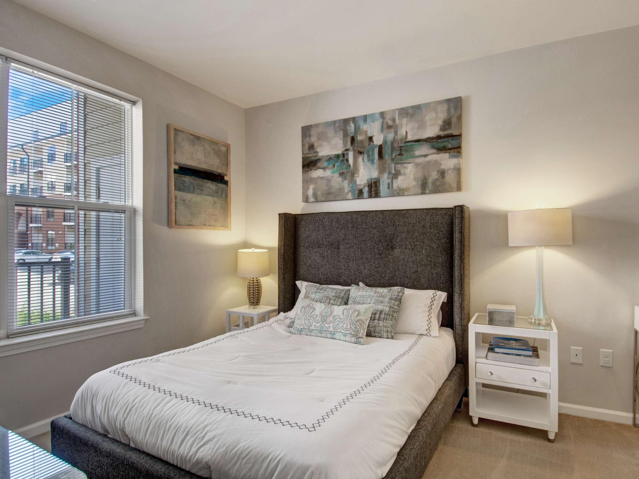Cozy bedroom with gray upholstered bed, neutral decor, and two windows providing exterior views.