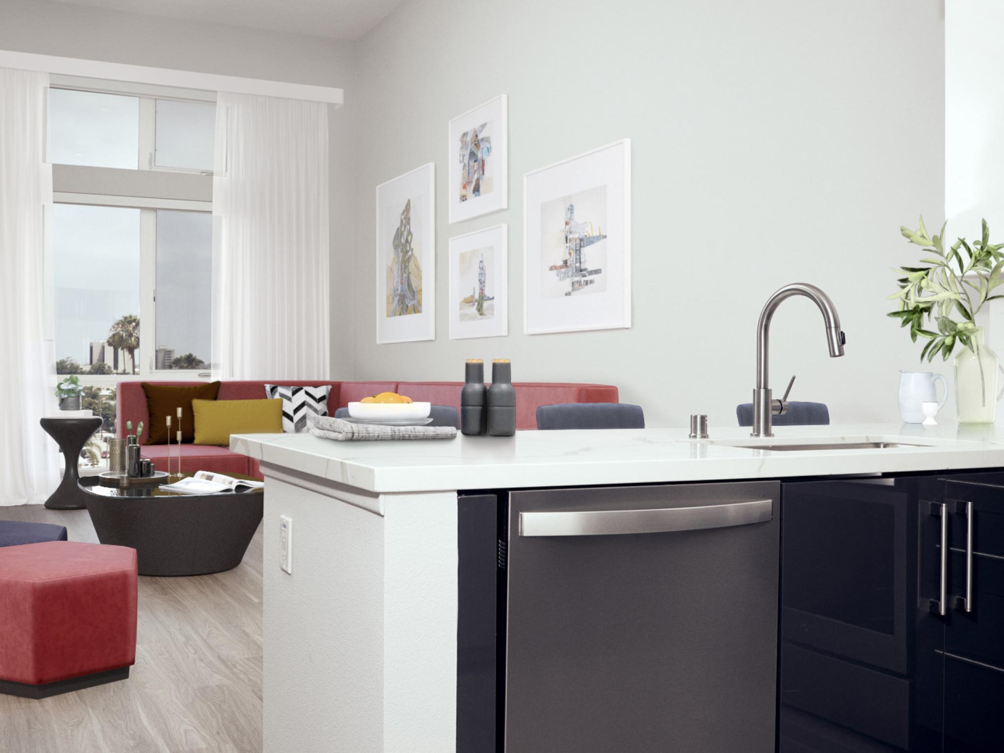 Modern kitchen island with stainless steel appliances and a gooseneck faucet, opening into a stylish living area with a red sectional, geometric ottomans, and framed wall art at The Linden.
