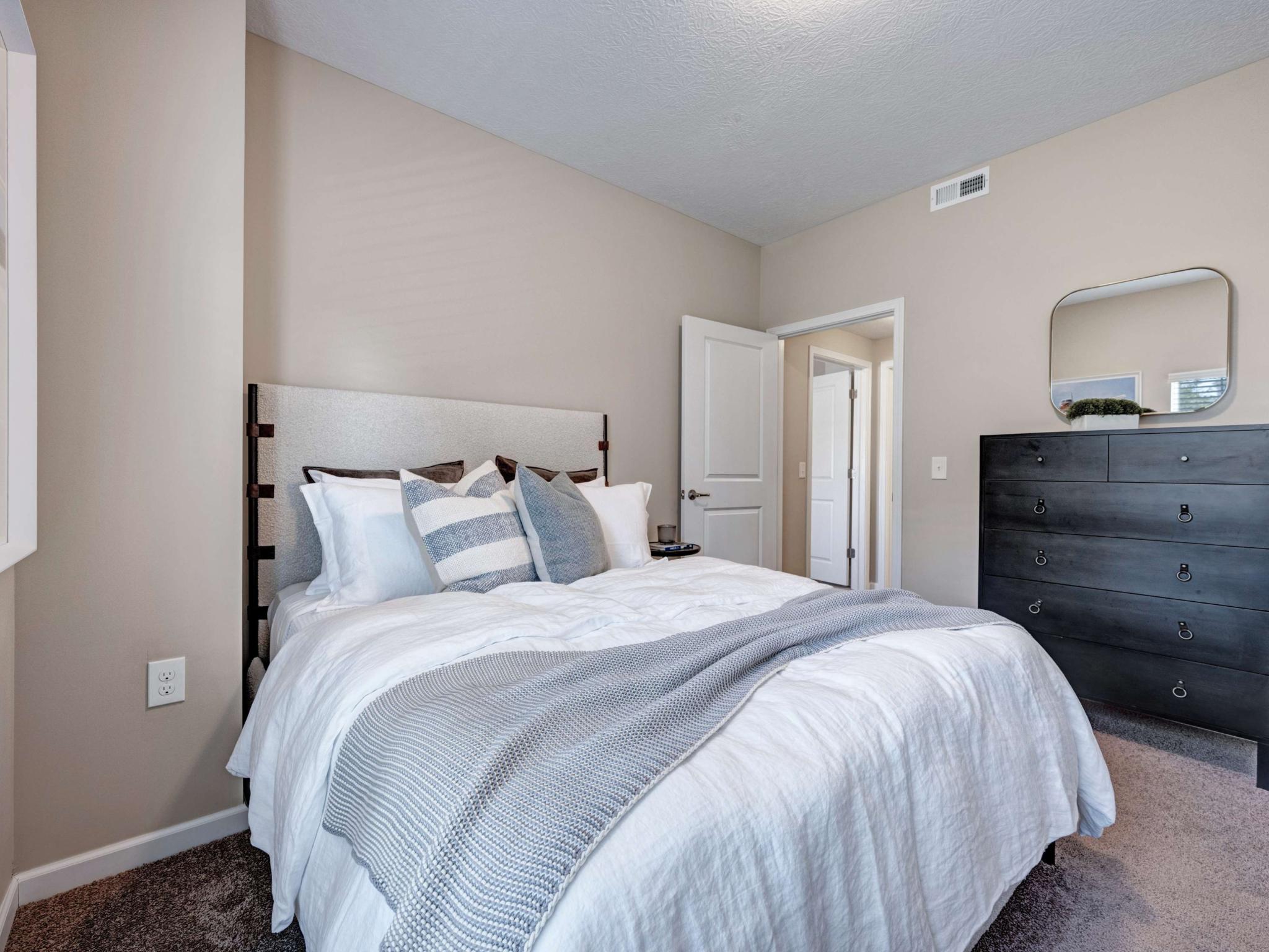 Comfortable bedroom with a neatly made bed, soft lighting, neutral walls, and a black dresser with a rounded mirror.