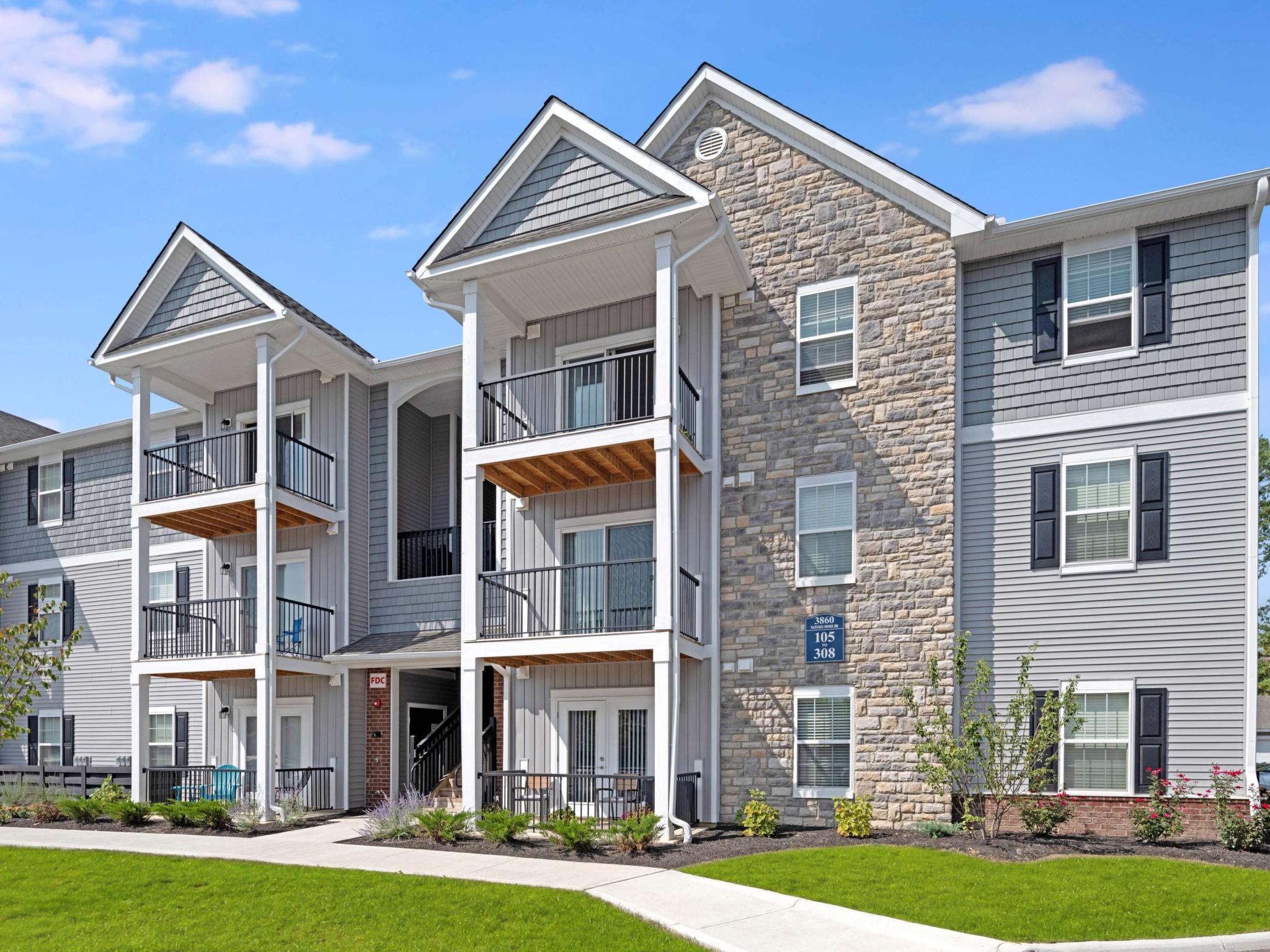 Three-story apartment building with gabled roofs, stone and siding exterior, and balconies overlooking the landscaped grounds.