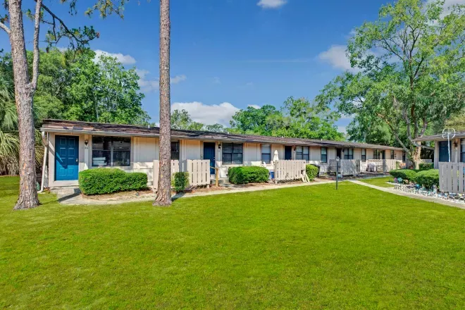 A row of single-story residential units with white picket fences and green lawns, surrounded by tall pine trees.