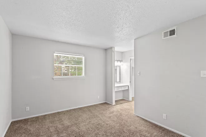 Empty bedroom with light gray walls, brown carpet, and a window. A vanity is visible through a doorway.