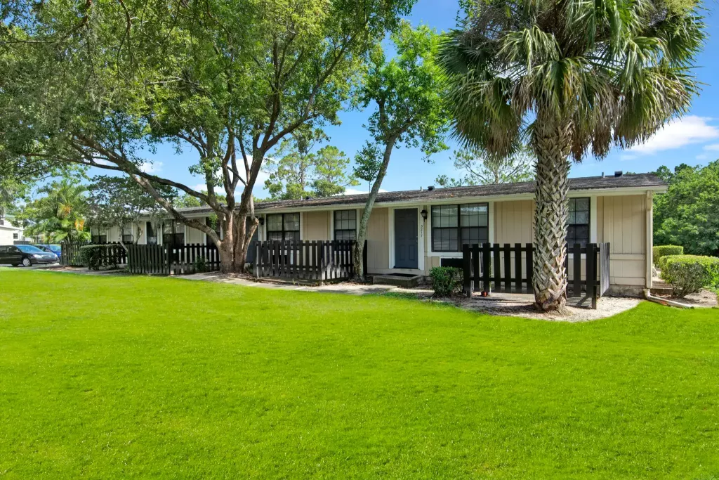 Exterior view of Branchwood Apartment Homes, featuring single-story residences with private fenced patios, surrounded by lush green lawns, mature trees, and a palm tree under a clear blue sky.