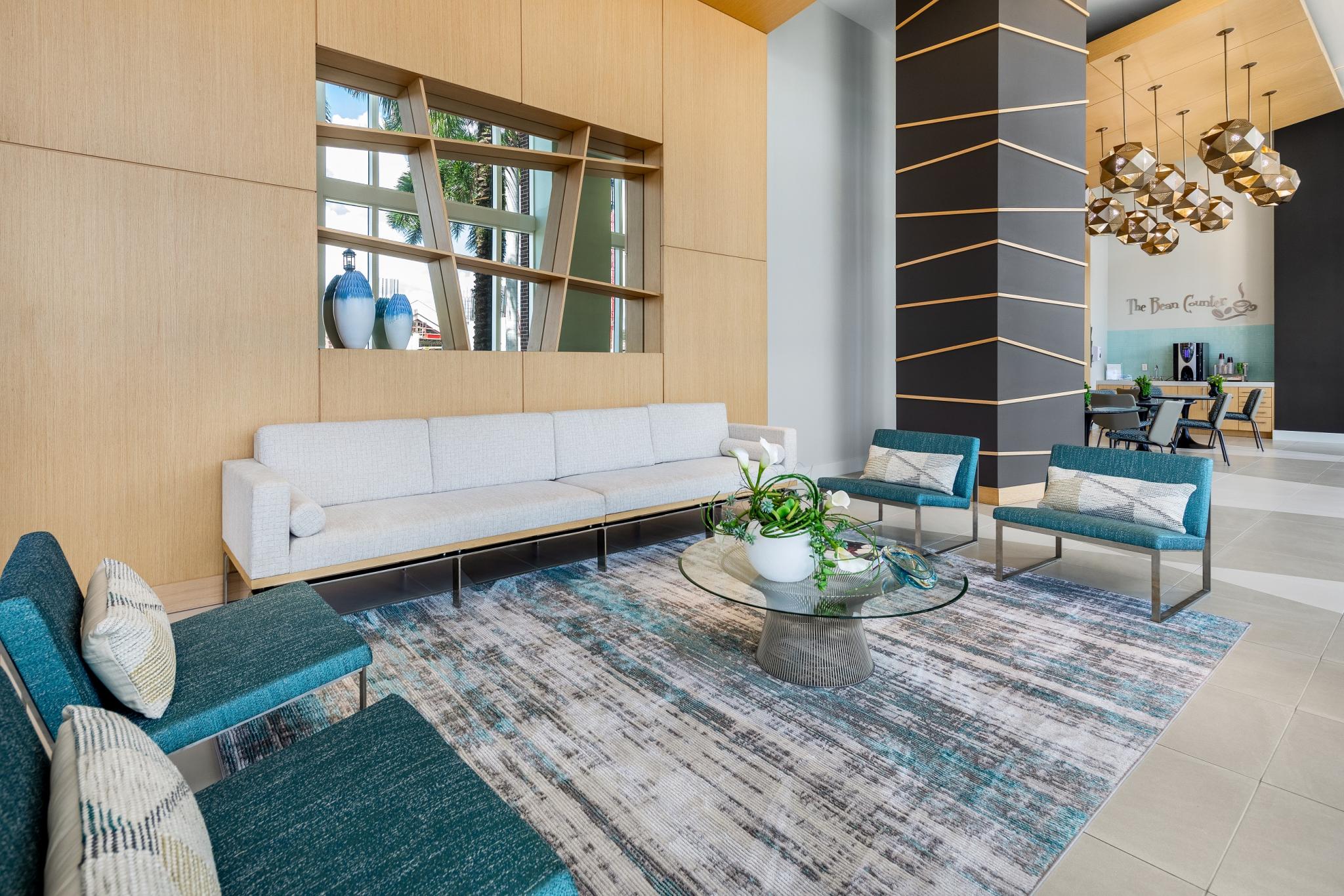 Modern apartment lobby at Midtown 24 in Plantation, Florida, featuring contemporary seating with teal accent chairs, a light gray sectional sofa, a round glass coffee table with a decorative plant, and a patterned area rug. The space includes wood-paneled walls, a geometric black column, and pendant lighting, with a coffee station labeled "The Bean Counter" in the background.