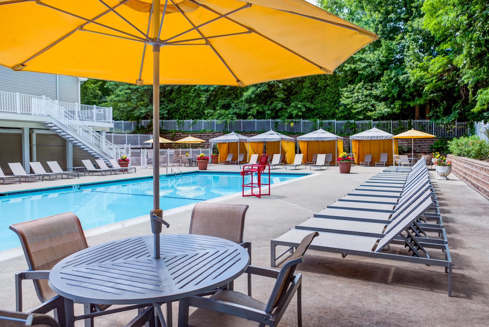 Accessible outdoor swimming pool with lounge chairs, shaded tables, yellow cabanas, and large umbrellas surrounded by greenery at a luxury apartment community.